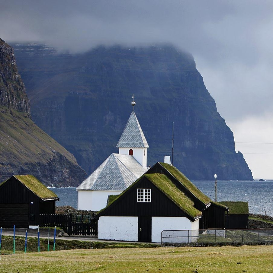 A white Faroese church and turf-roofed houses beneath a towering sea cliff